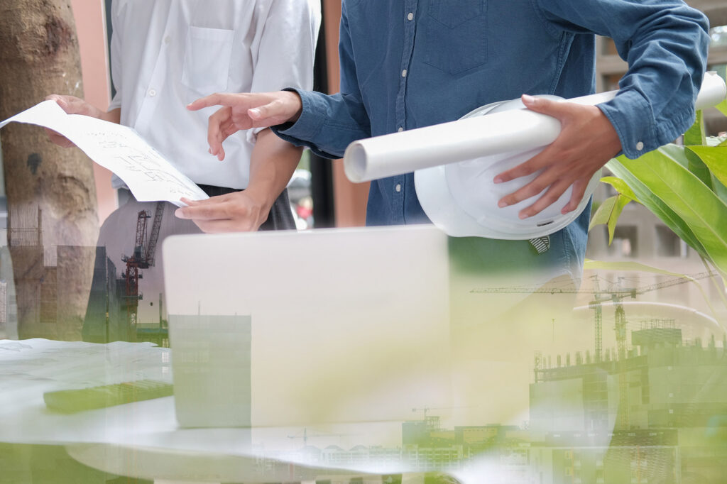 Specjaliści Giardini Polska analizują plany zielonej infrastruktury ESG, laptop, kask i zdjęcia z budowy BREEAM.